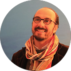 Homme souriant portant des lunettes et une écharpe, avec un fond bleu.
