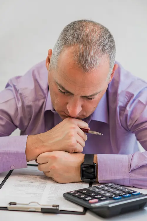 Homme pensif, penché sur un document, tenant un stylo et une calculatrice à portée de main, représentant les particuliers en dificulté pour leurs obligations fiscales